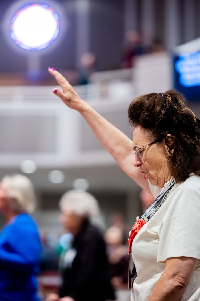 An elderly woman raises her hand in prayer inside a church setting.